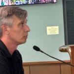 Side profile of a man with gray hair speaking into a microphone at a wooden lectern; a sign behind him reads 'No unauthorized personnel beyond this point'