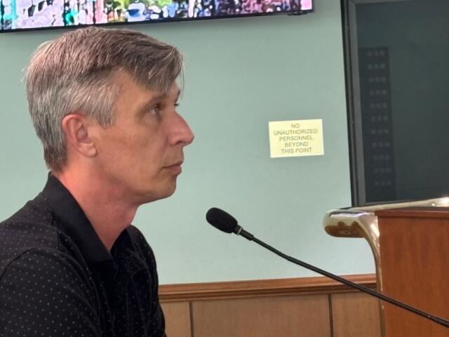 Side profile of a man with gray hair speaking into a microphone at a wooden lectern; a sign behind him reads 'No unauthorized personnel beyond this point'
