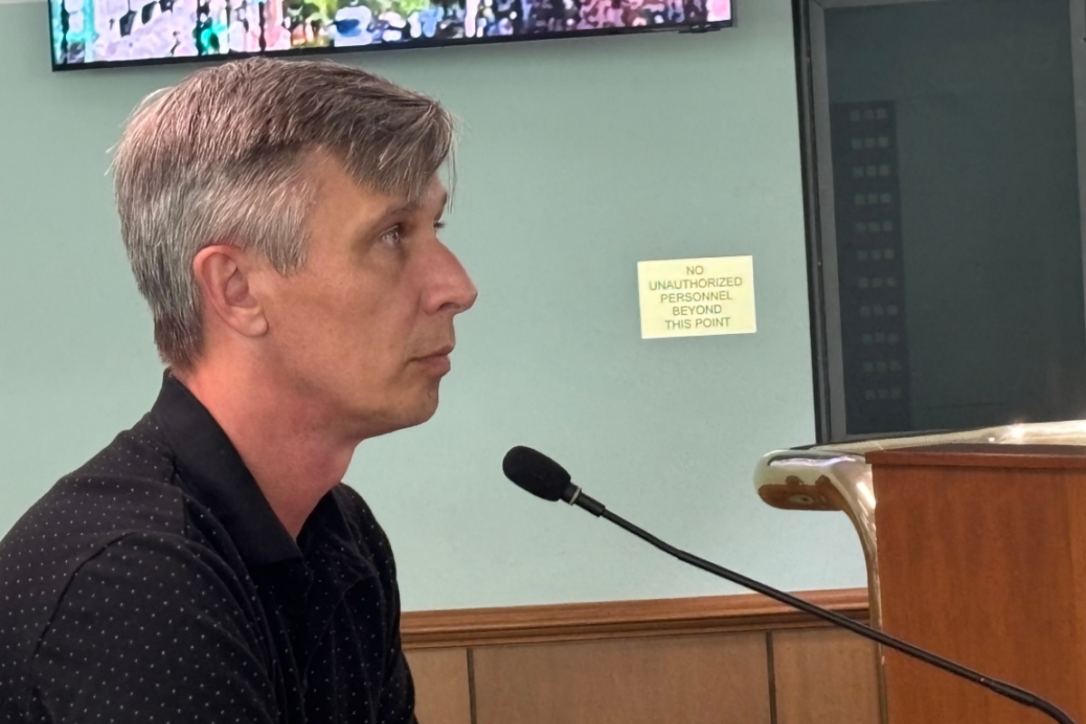 Side profile of a man with gray hair speaking into a microphone at a wooden lectern; a sign behind him reads 'No unauthorized personnel beyond this point'