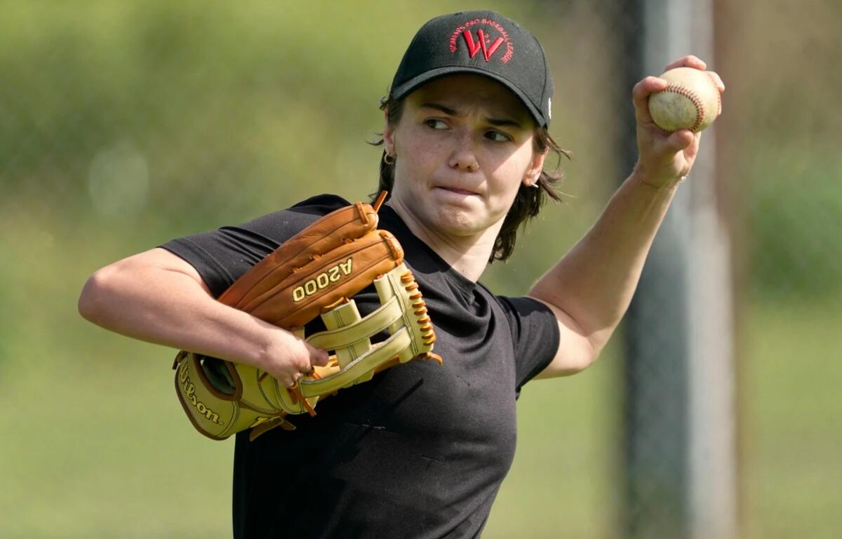 Female baseball pitcher in black shirt throwing a ball with her right hand, glove on left hand on a field.