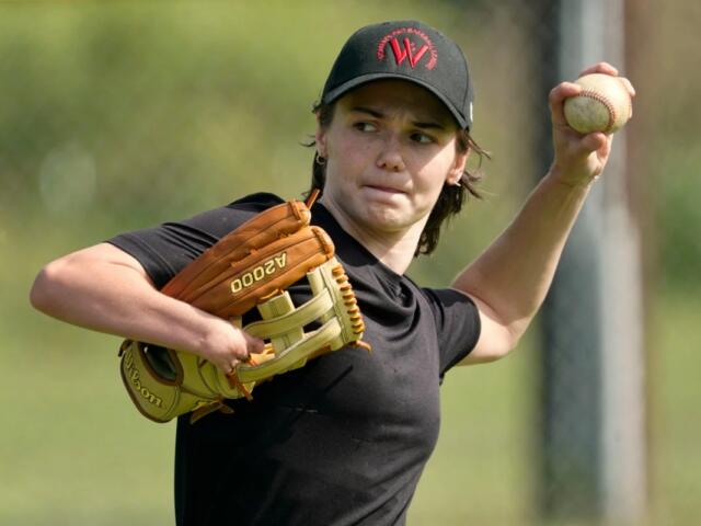 Female baseball pitcher in black shirt throwing a ball with her right hand, glove on left hand on a field.