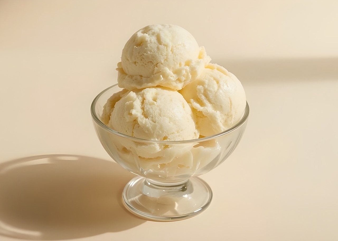 Three scoops of vanilla ice cream in a clear glass dessert bowl on a pale surface.