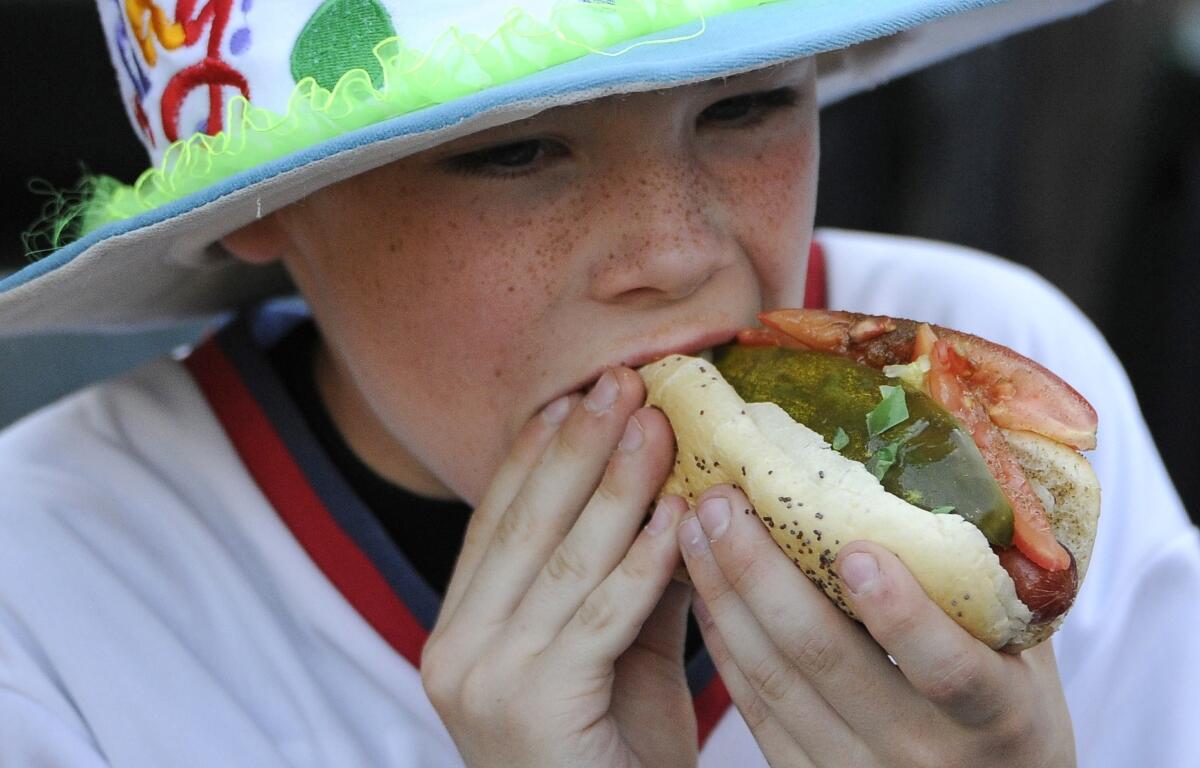 Child wearing a bright hat takes a big bite of a hot dog with pickle and vegetables outdoors, close-up view