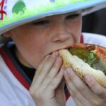 Child wearing a bright hat takes a big bite of a hot dog with pickle and vegetables outdoors, close-up view
