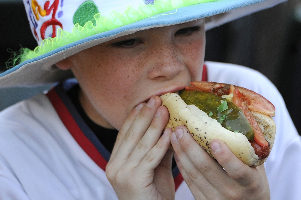 Child wearing a bright hat takes a big bite of a hot dog with pickle and vegetables outdoors, close-up view