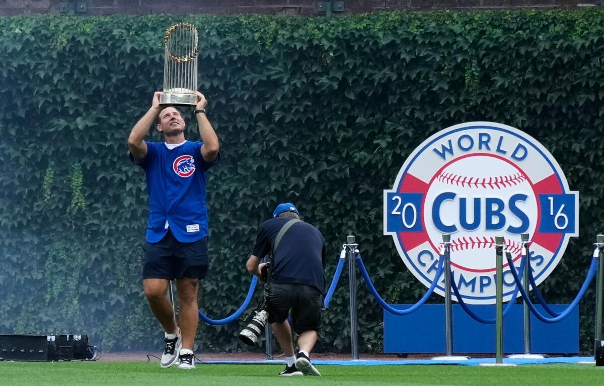 Cubs player lifts the World Series trophy above his head on the field, with a photographer nearby and a 2016 Cubs Champions display behind him.