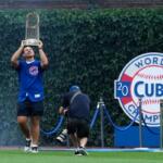 Cubs player lifts the World Series trophy above his head on the field, with a photographer nearby and a 2016 Cubs Champions display behind him.