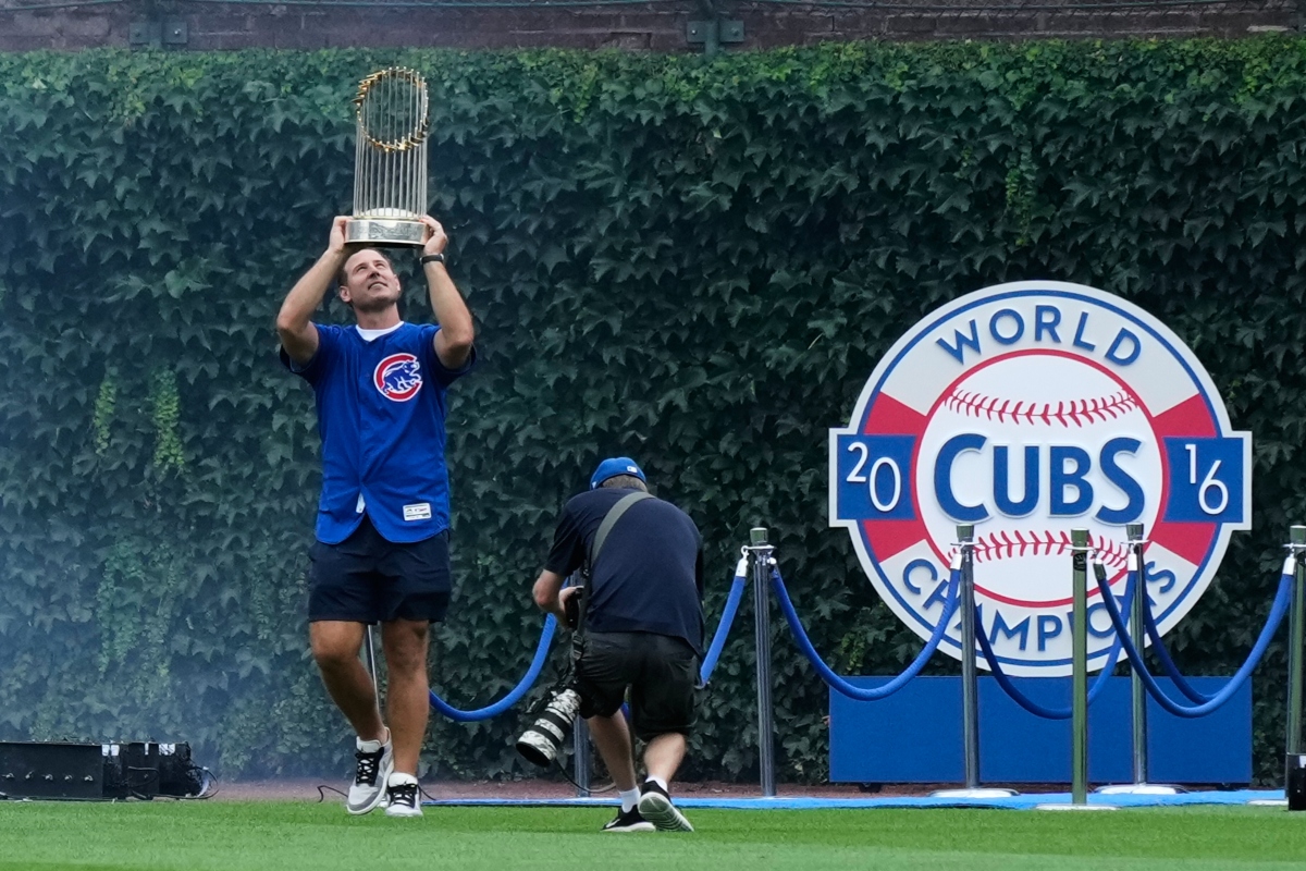 Cubs player lifts the World Series trophy above his head on the field, with a photographer nearby and a 2016 Cubs Champions display behind him.