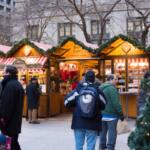 Outdoor Christmas market stalls with striped awnings and shoppers in winter coats at dusk, lit by festive lights.