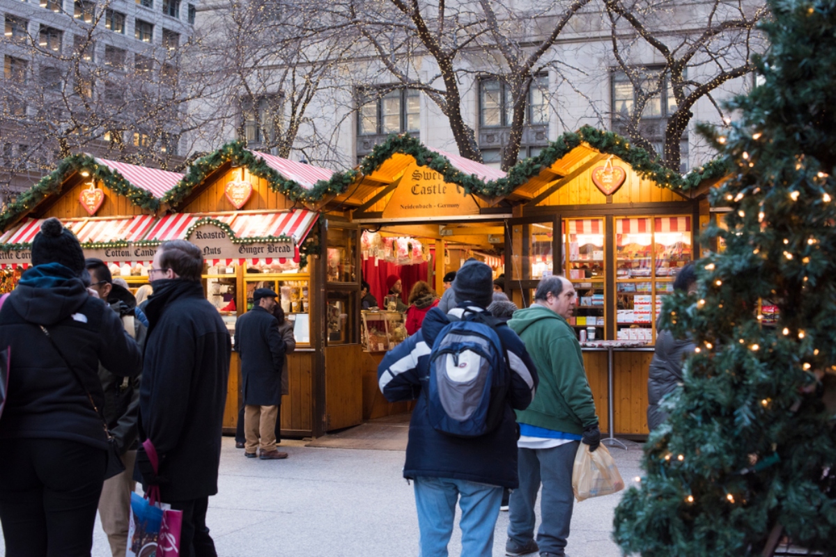 Outdoor Christmas market stalls with striped awnings and shoppers in winter coats at dusk, lit by festive lights.