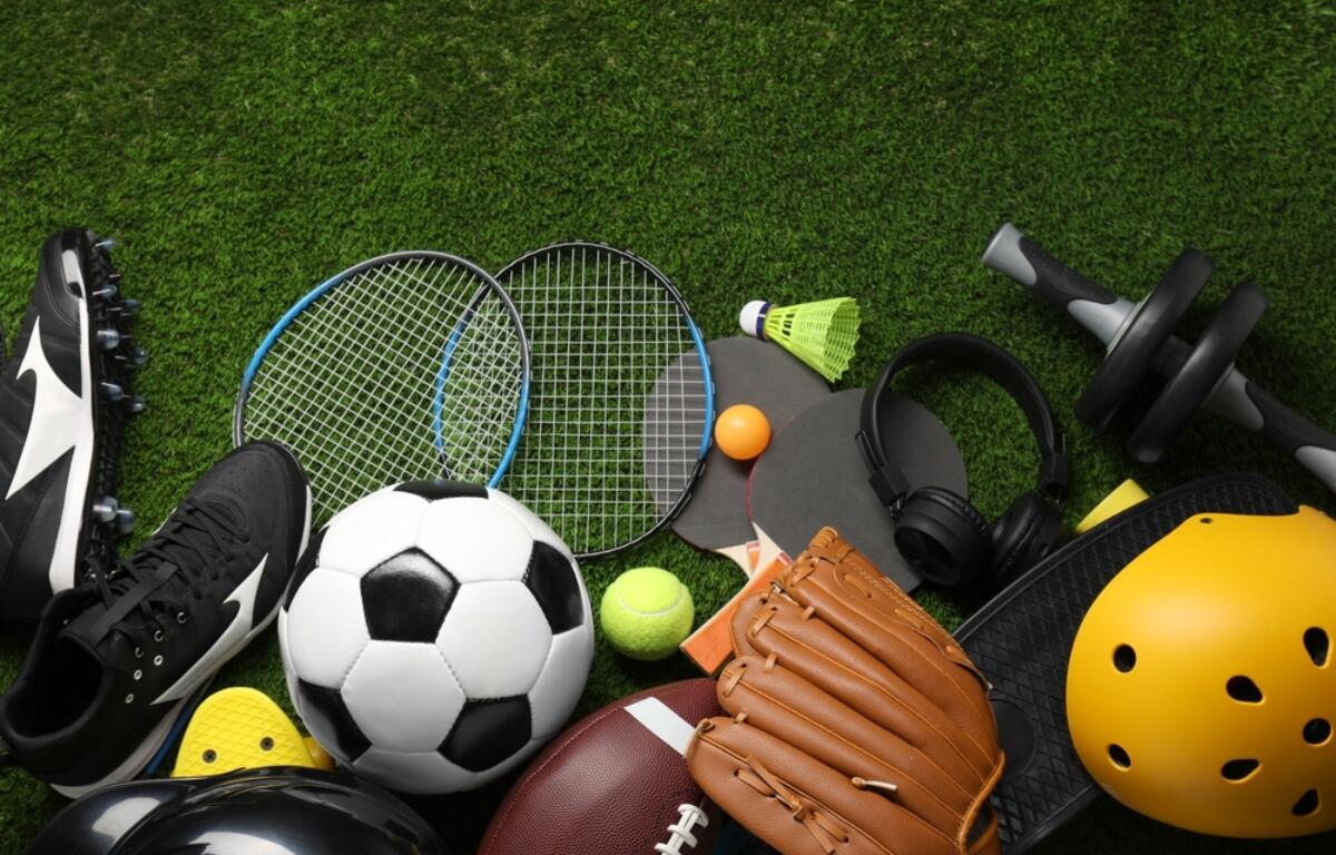 Assorted sports gear on green grass: soccer ball, tennis rackets, a yellow helmet, a baseball/football, gloves, shoes, a shuttlecock, and headphones scattered together.
