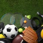 Assorted sports gear on green grass: soccer ball, tennis rackets, a yellow helmet, a baseball/football, gloves, shoes, a shuttlecock, and headphones scattered together.
