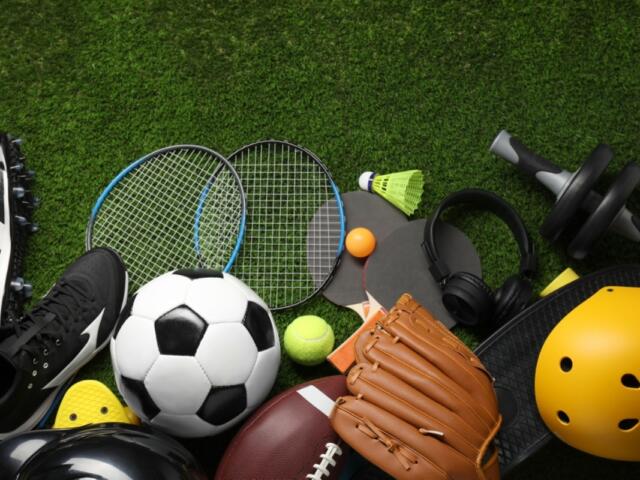 Assorted sports gear on green grass: soccer ball, tennis rackets, a yellow helmet, a baseball/football, gloves, shoes, a shuttlecock, and headphones scattered together.