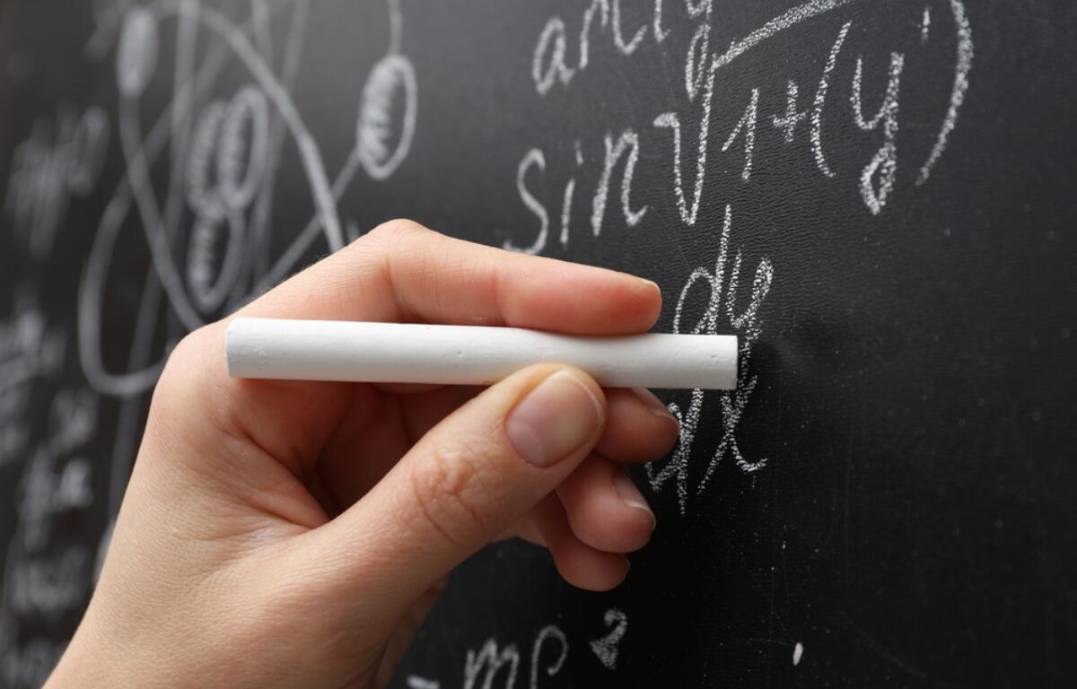 Close-up of a hand holding white chalk against a chalkboard filled with algebraic equations and diagrams.
