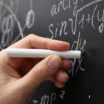 Close-up of a hand holding white chalk against a chalkboard filled with algebraic equations and diagrams.