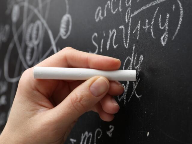 Close-up of a hand holding white chalk against a chalkboard filled with algebraic equations and diagrams.