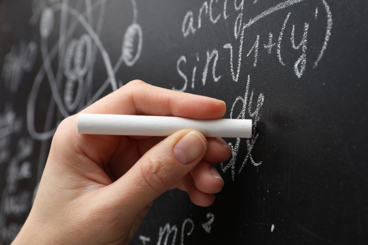 Close-up of a hand holding white chalk against a chalkboard filled with algebraic equations and diagrams.