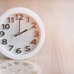 White round analog clock on a light wooden surface, showing almost twelve o'clock.