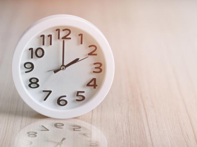 White round analog clock on a light wooden surface, showing almost twelve o'clock.