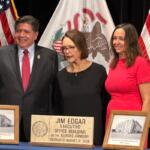Governor JB Pritzker, widow Brenda Edgar, and daughter Elizabeth, pose in front of plaques and a hand-made sign to be placed at the Illinois Armory, now the Jim Edgar Executive Office Building.
