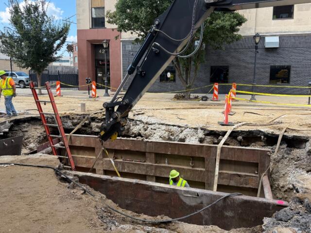 A City Water, Light, and Power employee works in the hole opened up in the road at the site of a water main break, while another employee is in the road.
