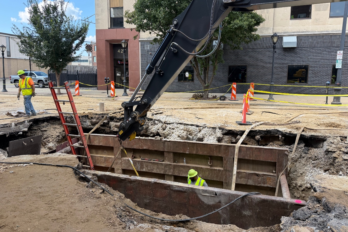 A City Water, Light, and Power employee works in the hole opened up in the road at the site of a water main break, while another employee is in the road.