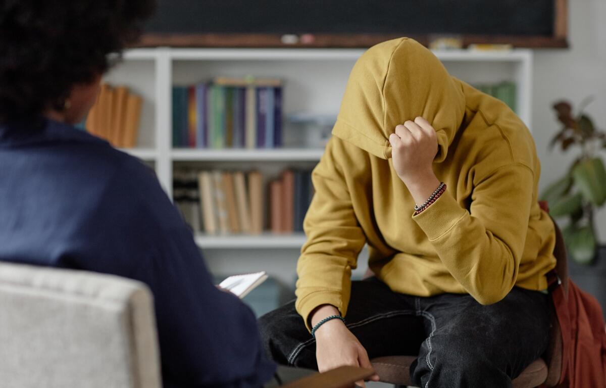 Portrait of distressed teenage boy sharing mental health troubles with therapist while sitting on chair and hiding face during one on one session copy space