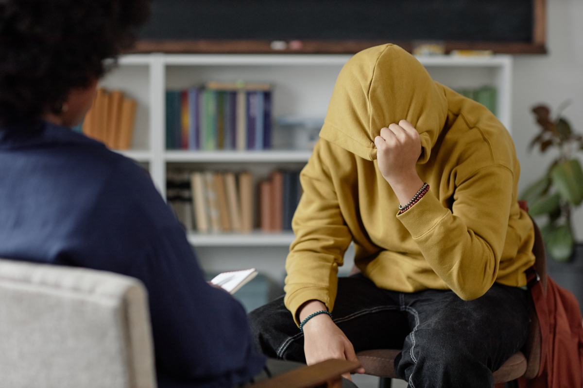 Portrait of distressed teenage boy sharing mental health troubles with therapist while sitting on chair and hiding face during one on one session copy space