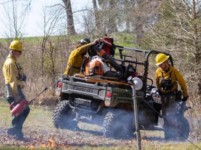 Left: SIPBA members unload a UTV March 20, 2026, during an interagency burn in Golconda, Illinois. Members pay $50 annually and are required to participate in one burn. In exchange, their land is also burned. Right: Pen Johnson secures a rake on Meredith Brown’s backpack March 20, 2026, during an interagency burn in Golconda, Illinois. Johnson and Brown are members of the Illinois chapter of The Nature Conservancy, a global residency program that aims to conserve lands and waters. (Photos by Carly Gist for the Saluki Local Reporting Lab)