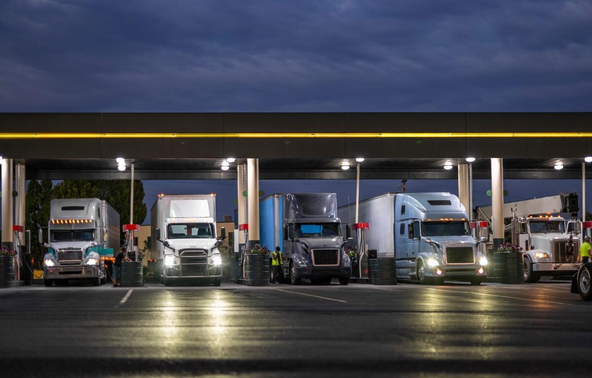 Industrial carriers big rig semi trucks with semi trailers fill the tanks with diesel fuel standing on the truck stop gas station with lights at night to be ready to continue the route by morning.