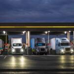 Industrial carriers big rig semi trucks with semi trailers fill the tanks with diesel fuel standing on the truck stop gas station with lights at night to be ready to continue the route by morning.