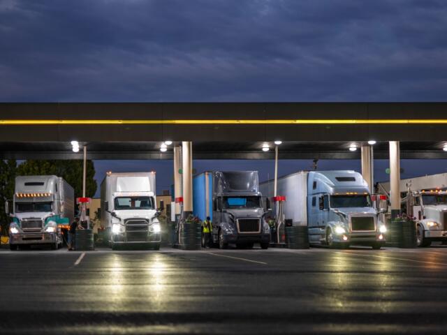 Industrial carriers big rig semi trucks with semi trailers fill the tanks with diesel fuel standing on the truck stop gas station with lights at night to be ready to continue the route by morning.