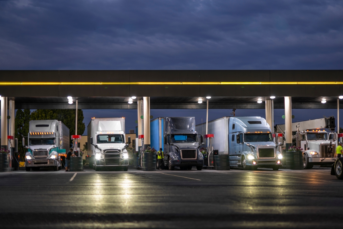 Industrial carriers big rig semi trucks with semi trailers fill the tanks with diesel fuel standing on the truck stop gas station with lights at night to be ready to continue the route by morning.