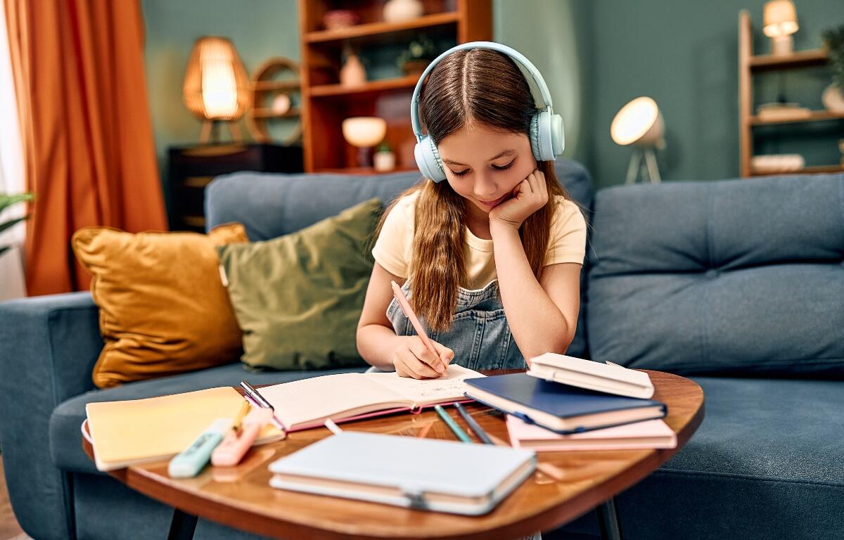 Young girl with headphones studying at a coffee table in a cozy living room, writing in a notebook surrounded by books and stationery.