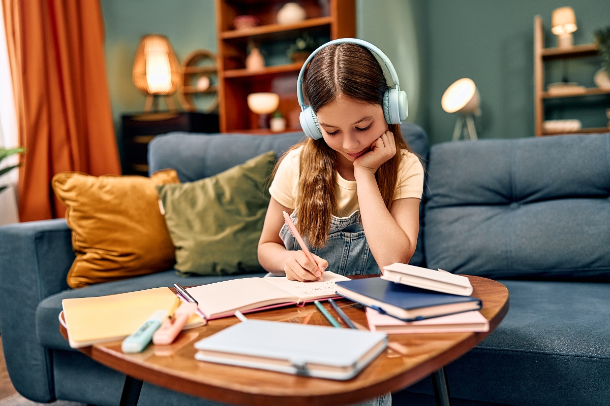 Young girl with headphones studying at a coffee table in a cozy living room, writing in a notebook surrounded by books and stationery.